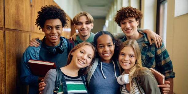 Six smiling teens holding books together in a school hallway